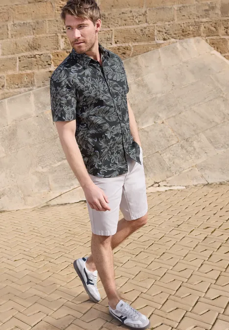Ein junger Mann in gemustertem Hemd, weißen Shorts und Sneakers steht entspannt auf einem gepflasterten Weg.