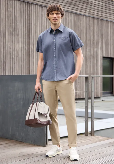 Man in donkerblauw overhemd, beige broek en sneakers, met tas voor een houten gevel.