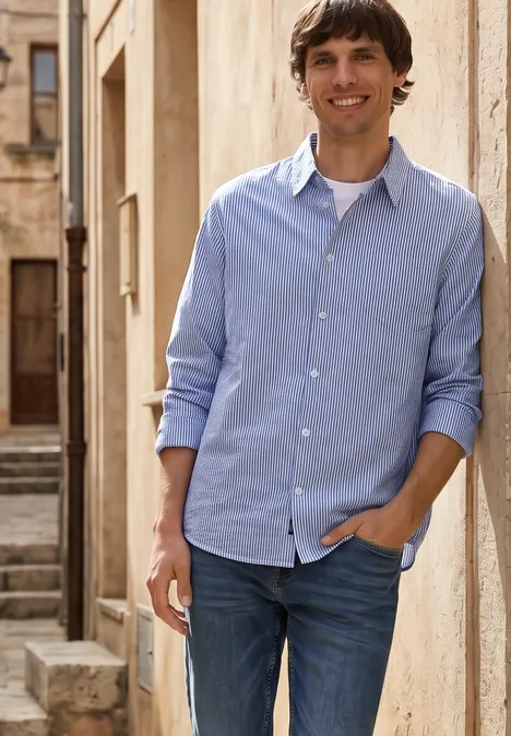 Overhemd met lange mouwen in seersucker stof bright white Een man staat voor een oude gevel, draagt een lichtblauw overhemd en jeans, met een hand in zijn zak.