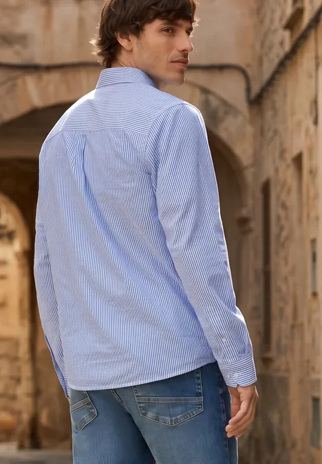 Overhemd met lange mouwen in seersucker stof bright white Een man met blauwe blouse en spijkerbroek, van achteren gezien in een lichtstenen gewelfgang.