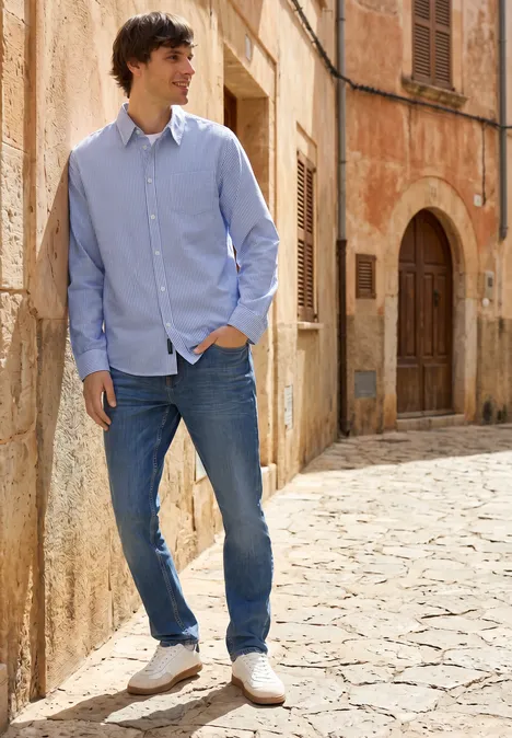 Overhemd met lange mouwen in seersucker stof bright white Een man in een lichtblauw overhemd, spijkerbroek en witte sneakers leunt ontspannen tegen een stenen muur in een steeg.