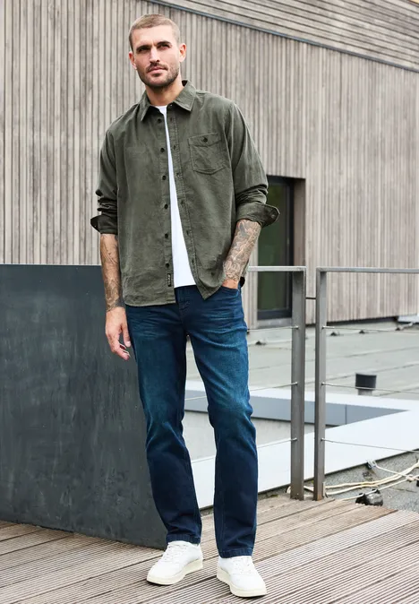 Effen corduroy overhemd met lange mouwen light moss green Man in kaki overshirt, wit T-shirt, blauwe jeans en witte sneakers op een dakterras.