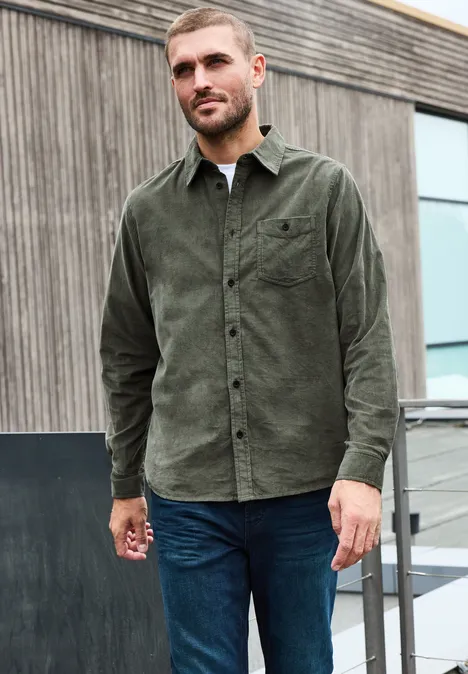Effen corduroy overhemd met lange mouwen light moss green Een man draagt een groen corduroy overhemd en blauwe jeans voor een modern houten gebouw.