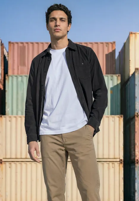 Ein Mann in weißem T-Shirt, schwarzer Jacke und khakifarbenen Hosen steht vor bunten Containern im Freien.