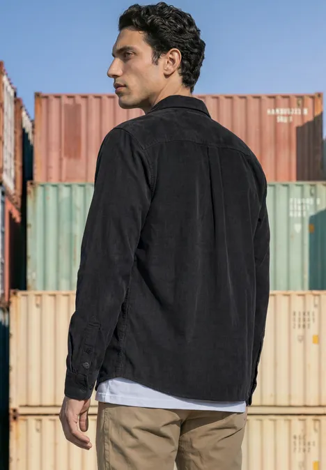 Effen corduroy overhemd met lange mouwen obsidian grey Een man in zwart overhemd staat voor gekleurde containers en kijkt opzij.