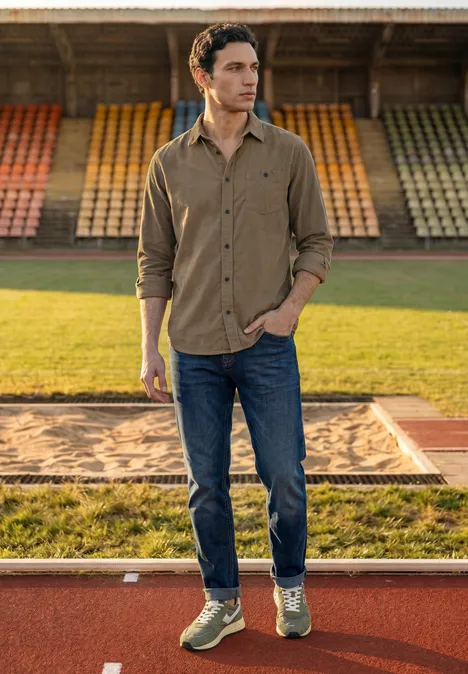 Effen corduroy overhemd met lange mouwen brass brown Een man in een bruine corduroy blouse, spijkerbroek en sneakers staat ontspannen op een baan.
