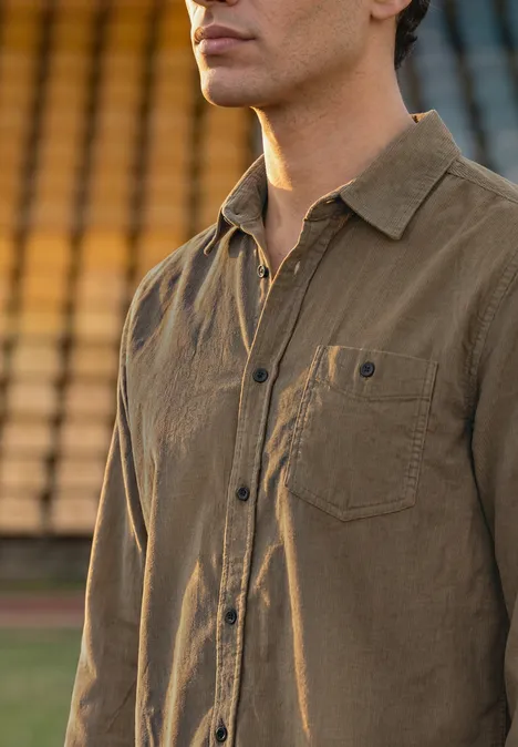Effen corduroy overhemd met lange mouwen brass brown Een man draagt een bruin overhemd, staand voor vage tribunes.