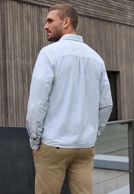 Overhemd met lange mouwen en streepjespatroon bright white Een man in licht overhemd en beige broek leunt ontspannen tegen een reling voor een houten gevel.