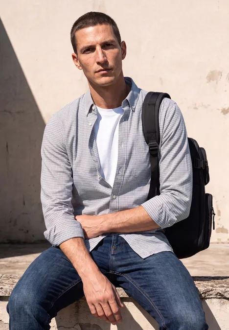 Langarm Popeline Hemd mit Print bright white Ein junger Mann mit Rucksack sitzt locker in Jeans und Hemd vor einer hellen Wand.