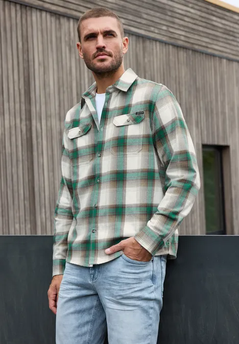 Overhemd met lange mouwen, borstzakken en ruitjespatroon bright white Man in groen-wit geruite blouse en spijkerbroek staat voor een houten gevel.