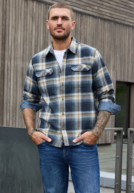 Langarm Hemd mit Brusttaschen und Karomuster dark ink blue Ein bärtiger Mann in karierter Flanelljacke und Jeans lehnt an einem Geländer vor einer Holzfassade.