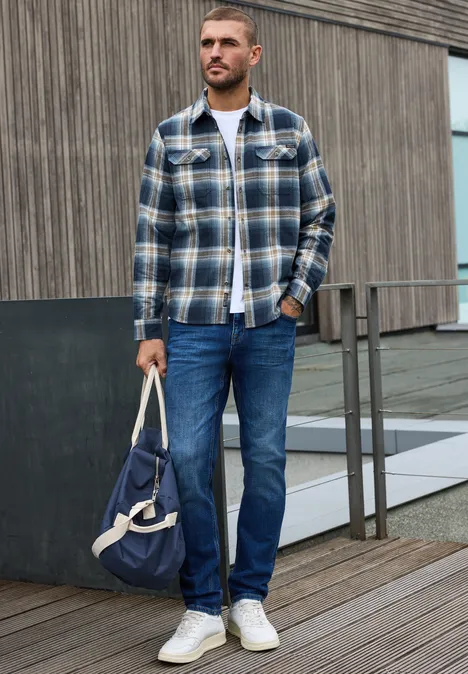 Langarm Hemd mit Brusttaschen und Karomuster dark ink blue Mann in kariertem Hemd, Jeans und weißen Sneakern hält draußen eine blaue Tasche.