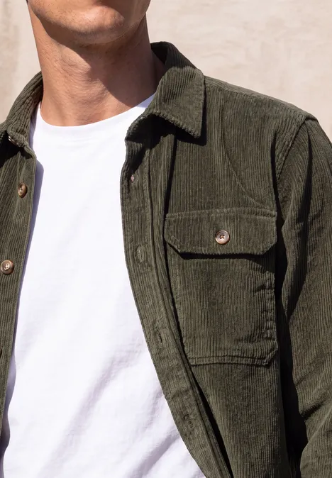 Ein Mann trägt eine olivgrüne Cordjacke über einem weißen T-Shirt vor hellem Hintergrund.