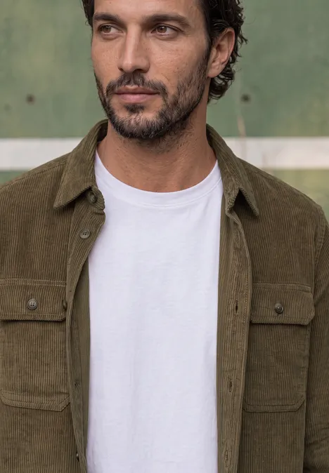 Ein bärtiger Mann trägt ein weißes T-Shirt und ein olivgrünes Cord-Hemd.