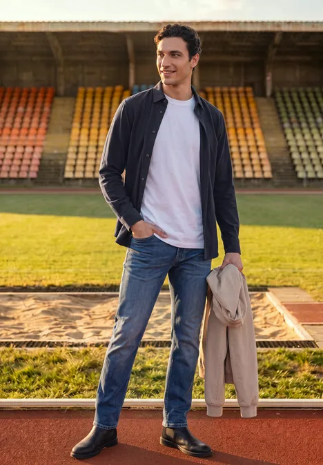 Ein lässig gekleideter Mann mit Jeans und Jacke steht neben einer Weitsprunggrube auf einer Sportanlage.