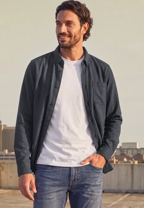 Langarm Twill Hemd mit Brusttasche basic black Mann steht auf Dachterrasse, trägt Jeans, weißes T-Shirt und dunkle Jacke, lächelt Richtung Kamera.