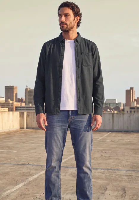 Langarm Twill Hemd mit Brusttasche basic black Ein Mann in Jacke, weißem T-Shirt und Jeans steht auf einem Parkdach vor Stadtkulisse.