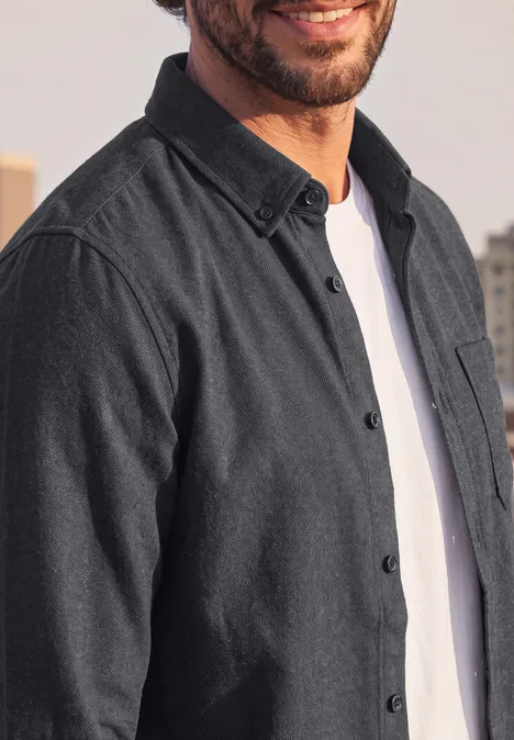 Langarm Twill Hemd mit Brusttasche basic black Ein Mann trägt ein dunkelgraues Hemd über einem weißen T-Shirt, nur Oberkörper und Lächeln sichtbar.
