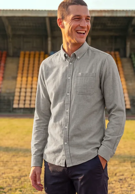 Chemise à manches longues en sergé avec poche poitrine antique beige Ein freundlicher Mann in grauem Hemd lächelt vor leeren Tribünen im Stadion.