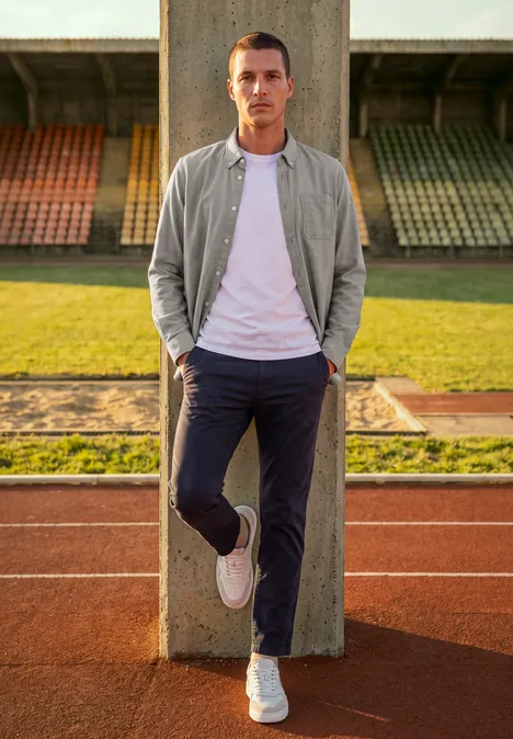Chemise à manches longues en sergé avec poche poitrine antique beige Ein Mann lehnt entspannt an einer Betonstütze auf einer Laufbahn vor Stadionrängen.