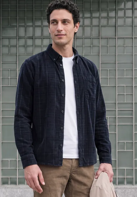 Ein junger Mann steht vor einer Glasfliesenwand, trägt weißes T-Shirt, offenes kariertes Hemd und braune Hose.