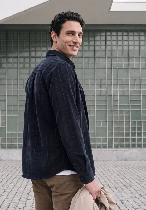 Ein junger Mann in Jeans und dunklem Hemd blickt über die Schulter vor einer Gitterwand.