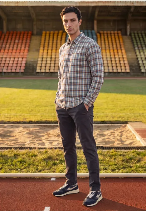 Chemise en flanelle à manches longues à carreaux raw copper red Ein Mann in kariertem Hemd, dunklen Hosen und Sneakern steht auf einer Laufbahn vor leeren Tribünen.