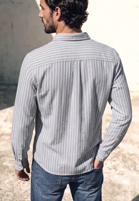 Chemise en flanelle à manches longues à rayures cement grey melange Un homme de dos, portant une chemise bleu clair rayée et un jean, se tient devant un mur gris.