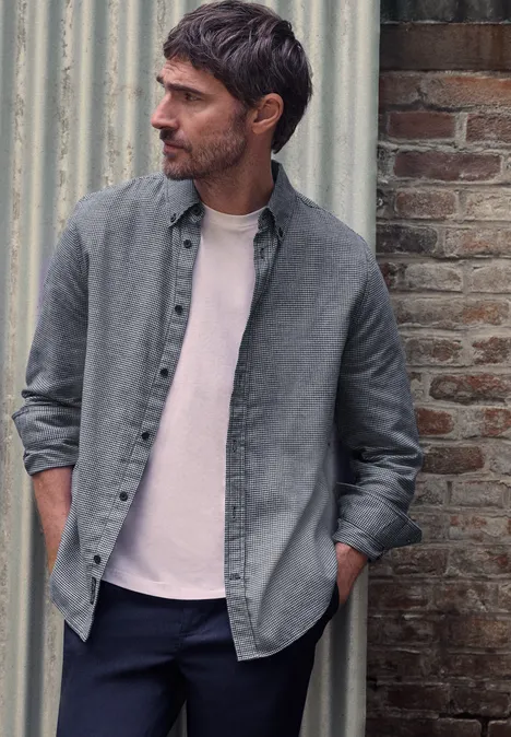 Ein bärtiger Mann in grauem Hemd und rosa T-Shirt lehnt an einer Wand und schaut zur Seite.