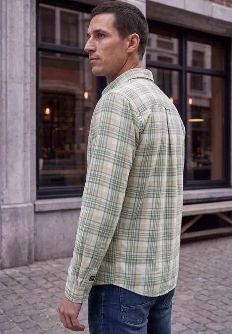 Een man in profiel draagt een lichtgroen geruit hemd en jeans, staand voor een gebouw in de stad.
