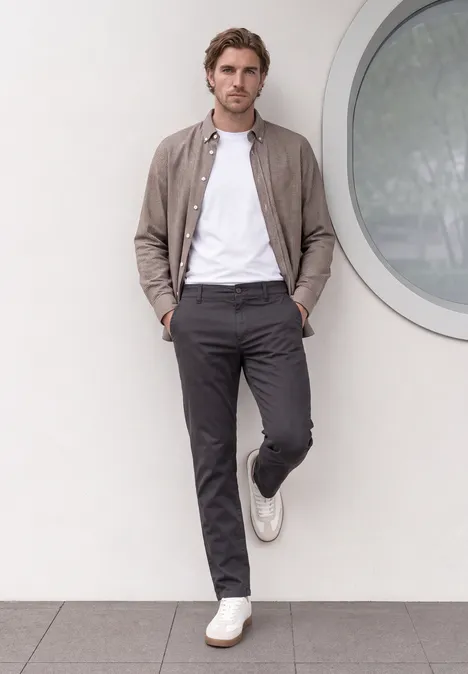 Ein Mann lehnt entspannt an einer Wand, trägt weißes T-Shirt, graue Hose, braune Jacke und weiße Sneaker.