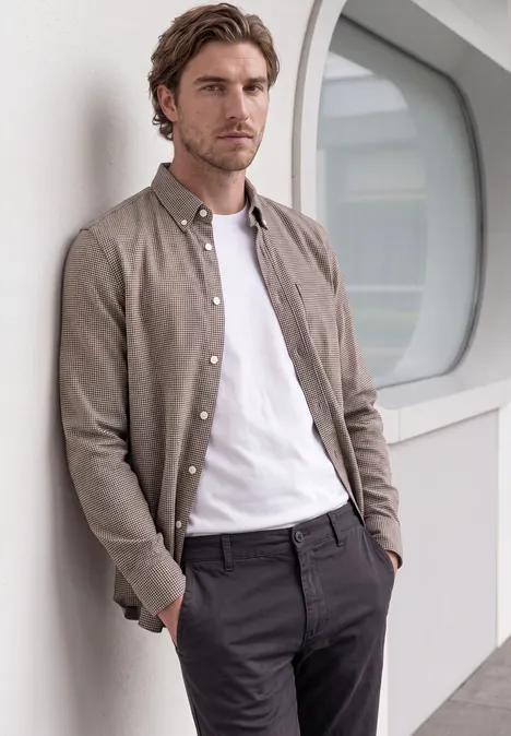 Ein Mann lehnt lässig an einer Wand, trägt ein weißes T-Shirt, ein braunes Hemd und eine dunkle Hose.
