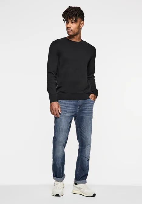 Homme en pull noir, jean et baskets blanches regarde sur le côté.