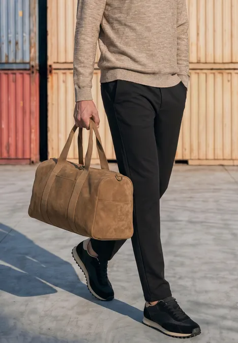 Eine Person trägt schwarze Hose und hält eine braune Reisetasche vor bunten Containern.