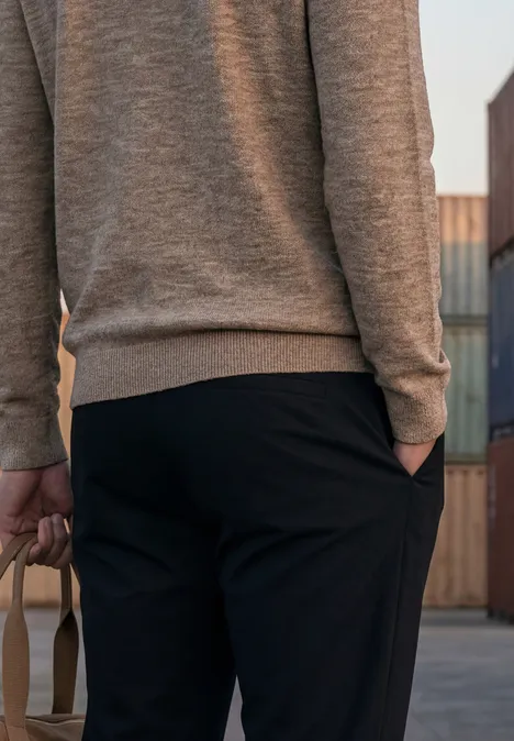 Rückansicht einer Person in grauem Pullover und schwarzer Hose, die eine Tasche hält, vor Containern.