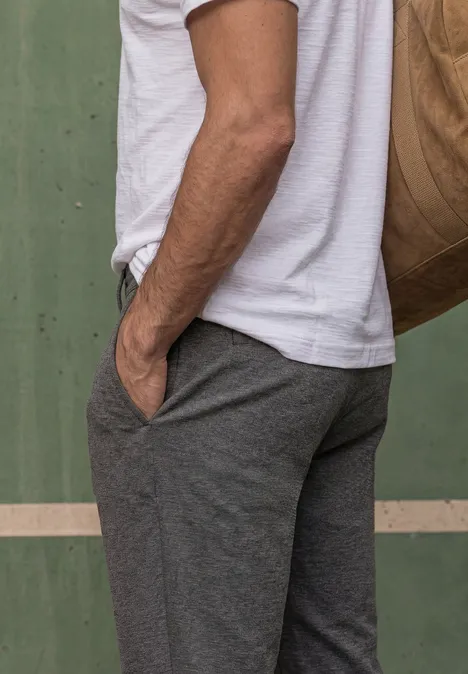 Ein Mann in grauer Hose und weißem T-Shirt steht seitlich, Hand in der Tasche, mit Umhängetasche.
