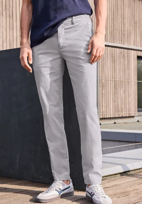 Ein Mann trägt graue Chino-Hose und Sneaker, nur Unterkörper, vor hölzerner Wand