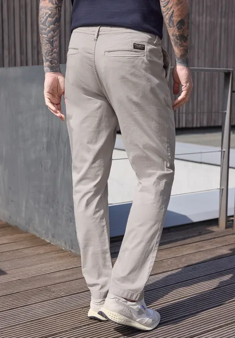 Un homme vu de dos porte un pantalon chino clair sur une terrasse en bois près d’une balustrade.