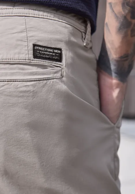 Gros plan sur un pantalon homme gris, poche arrière à écusson, main dans la poche latérale et tatouage au bras