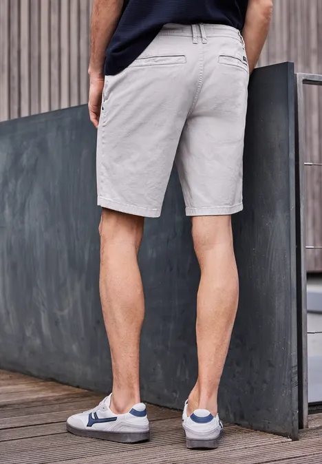 Ein Mann lehnt an einer Betonwand, trägt graue Shorts und weiße Sneaker.