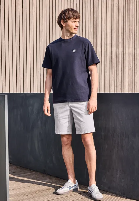 Ein Mann in dunkelblauem T-Shirt, hellgrauen Shorts und weißen Turnschuhen steht vor einer modernen Holzfassade.