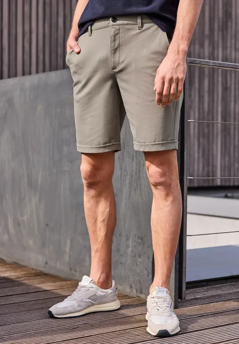 Ein Mann trägt beige Shorts, dunkles T-Shirt und weiße Sneaker, stehend vor einer modernen Holzfassade.