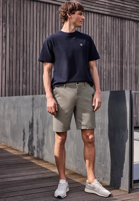 Ein Mann in schwarzem T-Shirt, grauen Shorts und weißen Sneakern steht auf einem Holzsteg vor einer grauen Wand.