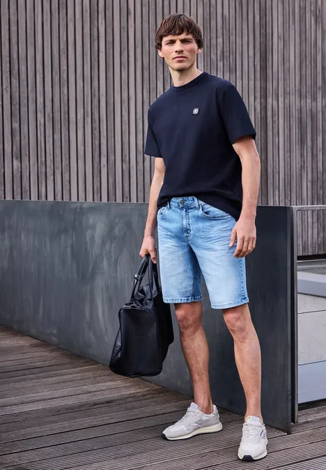 Ein Mann in lässigem Outfit: schwarzes T-Shirt, Jeans-Shorts und weiße Sneaker mit schwarzer Tasche auf Holzdeck