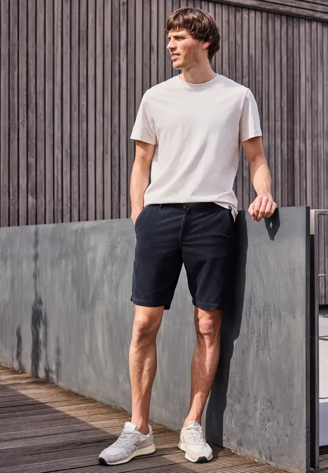 Ein Mann in weißem T-Shirt und schwarzen Shorts steht entspannt auf einem Holzsteg vor einer Fassadenwand.