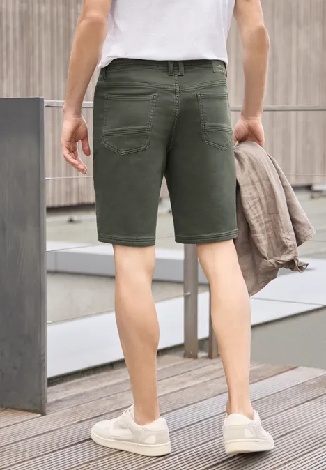 Man op terras gezien van achter, draagt olijfkleurige korte broek en witte sneakers, houdt jasje vast.