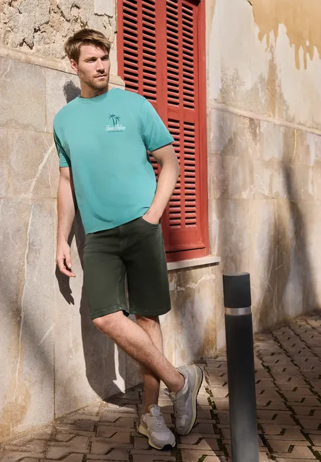 Een jonge man in een turquoise shirt en donkere shorts leunt ontspannen tegen een zonnige gevel met rode luiken.