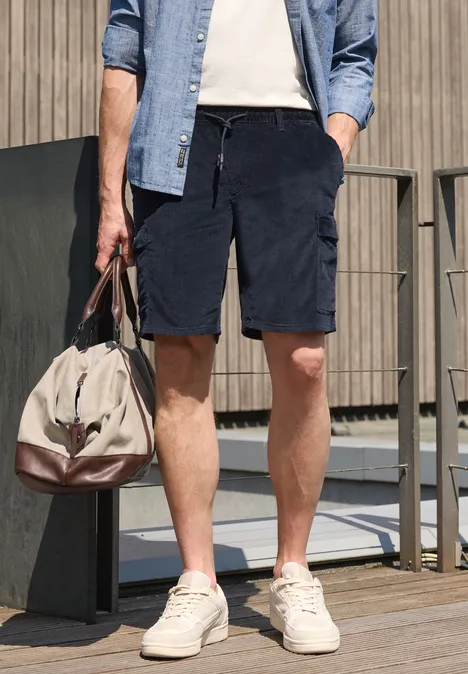 Homme en chemise en jean, T-shirt blanc, short bleu marine en velours côtelé, baskets et sac de voyage.