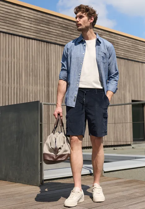 Un homme en chemise, t-shirt, short et baskets se tient dehors en tenant un sac de voyage.