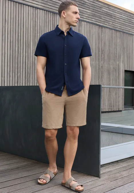 Un homme en polo bleu marine, short beige et sandales, pose devant une moderne façade en bois.